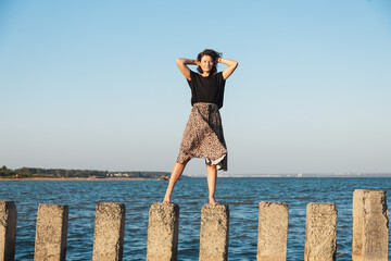 Young beautiful woman in a light casual dress walking on rocks on a sandy beach near a blue sky on a summer day, beautiful sea against a blue sky