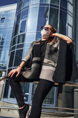 Young woman in protective mask standing outdoors near business building at sunny daytime. Conception of coronavirus