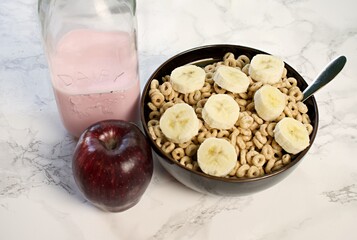 Close up on bowl of cereal with banana slices, spoon, red apple, and strawberry milk. 