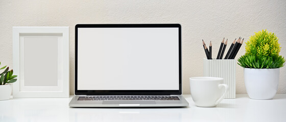 Stylish workspace with blank screen laptop, frame, tree, pencil and coffee cup on white background in office. Front view