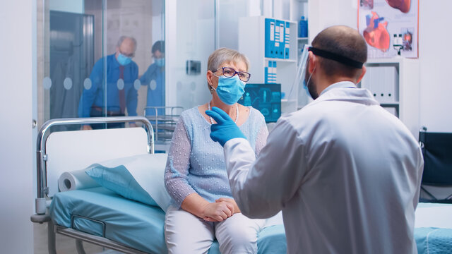 Old senior woman having a medical consultation during COVID-19 pandemic, wearing a mask and doctor in protective wear. Modern hospital or private clinic. Healthcare system