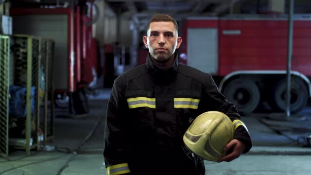 Firefighter video portrait wearing uniform and holding yellow protective helmet. Fire trucks in the background. Camera panning left.