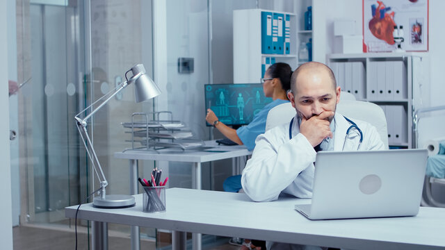 Man doctor typing on laptop in modern office with glass walls, medical staff and patients walking in hallway. Healthcare system clinic consultation from practitioner physician. Handheld shot - Powered by Adobe