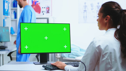 Physician writing diagnose on computer with green screen and assistant wearing blue uniform in the background. Medic in white coat working on monitor with chroma key in clinic cabinet to check patient