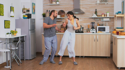 Joyful couple dancing and singing during breakfast in kitchen wearing pajamas. Carefree wife and...