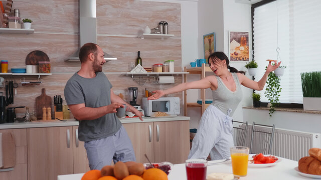 Married Couple Having Fun Dancing In Kitchen During Breakfast. Carefree Husband And Wife Laughing, Singing, Dancing Listening Musing, Living Happy And Worry Free. Positive People.