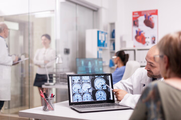 Radiologist pointing at brain ct scan on laptop screen during senior woman check up in hospital office. Mature doctor in white coat discussing with sick young woman in clinic corridor.