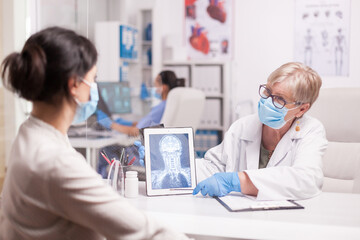 Obraz premium Lekarz z maską agasint covid-19, wskazujący na tablet pc z prześwietleniem mózgu podczas konsultacji z pacjentem.