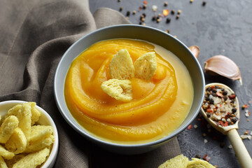 Pumpkin creamy soup with wheat croutons, garlic and spices in ceramic bowl with wooden spoon on grunge black background with linen napkin