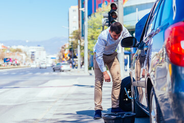 Obraz premium A sweaty tired young entrepreneur is wiping the sweat off his forehead while trying to change a flat tire on a beautiful sunny day