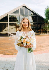Fototapeta premium Beautiful blonde bride holding a bouquet in her hands on the background of a modern building