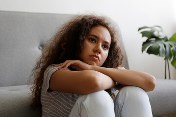 Naklejka premium Portrait of young beautiful black woman with depressed facial expression sitting on the couch touching her temples. Female in physical and emotional pain. Sad girl in her room. Background, copy space.
