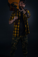 Tattooed man with a beard wearing a tartan yellow black shirt is  posing holding his guitar on his sholder and giving his middle finger