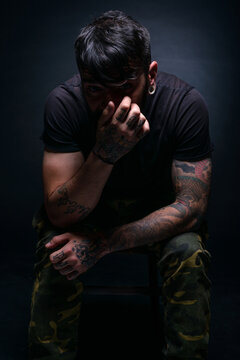 High contrast picture of a bearded male model sitting on a chair and covering his face with his tattooed hands