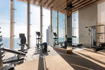 Exercise Bike and Treadmill Equipment in Modern Gym with views of Sea and Sky in Background of Large Glass Windows, The sun's rays penetrated the big window into the luxury gym. - obrazy, fototapety, plakaty