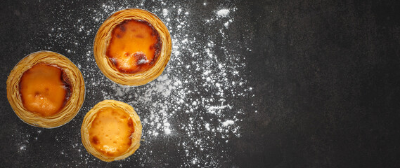 top view of three tasteful egg tarts and wheat flour on dark grey texture background with copy...