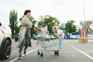 Happy mother and her daughters are having fun with a shopping cart on a parking lot beside a...