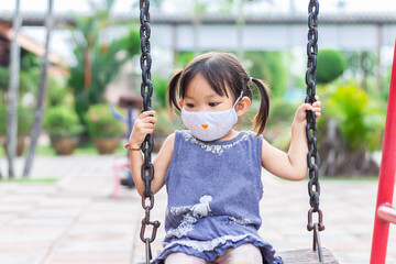 Happy​ Asian​ child​ girl​ wearing​ a​ fabric​ face​ mask​ when​ she​ playing​ a​ toy​ at​ the​ playground.​ Social​ Distance.​