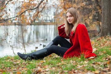 Obraz premium Portrait of a blonde girl in a red coat sitting on the shore of a lake in an autumn park