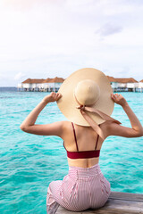 Girl on the wooden jetty looking to the ocean. Beautiful woman on wooden pier by the sea outdoors holding her hat. Back view of a young girl sitting on a pier with sea and sky in background.