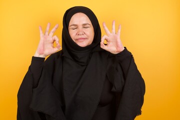 Beautiful middle aged muslim woman wearing black hijab over yellow background relax and smiling with eyes closed doing meditation gesture with fingers. Yoga concept.