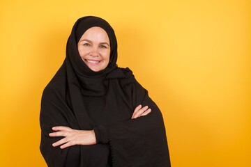 Beautiful middle aged muslim woman wearing black hijab over yellow background happy face smiling with crossed arms looking at the camera. Positive person.