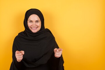 Middle aged muslim woman wearing black hijab over yellow background pointing fingers to camera with...