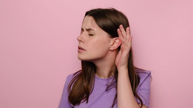 Confused young woman having hearing problems, asking to say louder, difficult to listen quiet talk, dressed in purple t-shirt, misunderstanding in communication, isolated on pink studio background