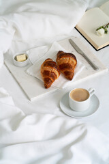 Croissants on the napkin, knife and butter on the marble tray with a cup of coffee and book on the white bed with blanket. Homemade breakfast in bed.