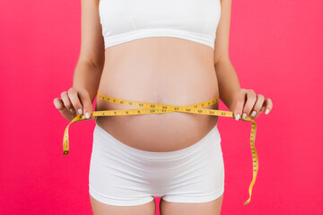 Close up of pregnant woman in white underwear measuring her growing belly at pink background. Expecting a baby. Inch measurement. Copy space