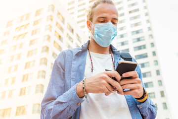 caucasian young man wearing a blue surgical mask walking in the city while using a phone