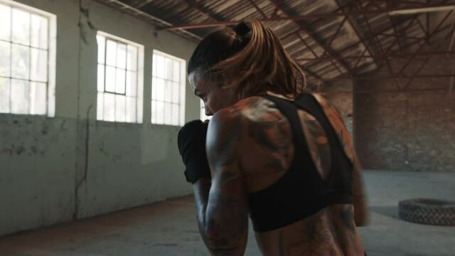 Female doing shadow boxing in epmty factory shade. Tattooed woman in sportswear practicing her punches at an abandoned warehouse.
