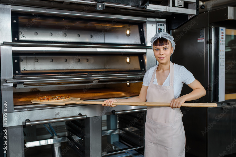 Wall mural young caucasian woman baker is holding a wood peel with fresh pizza near an oven at a baking manufac