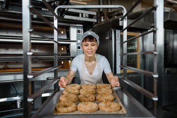 Close up of young caucasian woman baker is holding a tray with fresh bagels and looking to them at...