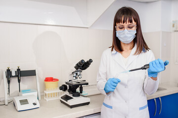Young woman doctor laboratory assistant is using micropipette test tubes near a medical microscope and urine strip analyser.