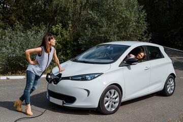 Obraz na płótnie Canvas Two happy young beautiful women are talking to each other until their electric car is charging at the charging station situated in the forest.