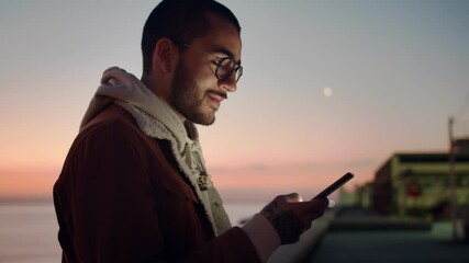 Side portrait of handsome confident young guy in glasses staying online outdoors sunrise, cheerful millennial man influencer using smartphone device smiling connected in social media network - Powered by Adobe