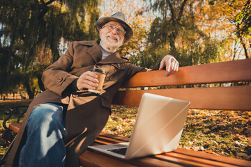 Photo of white grey hair positive old man enjoy autumn nature outside park rest relax sit bench with takeaway coffee mug use laptop look sunshine wear coat jacket hat cap headwear