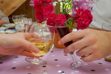 Glasses with red and white wine in the hands of a man and a woman are clinking against the background of a festive table and flowers.