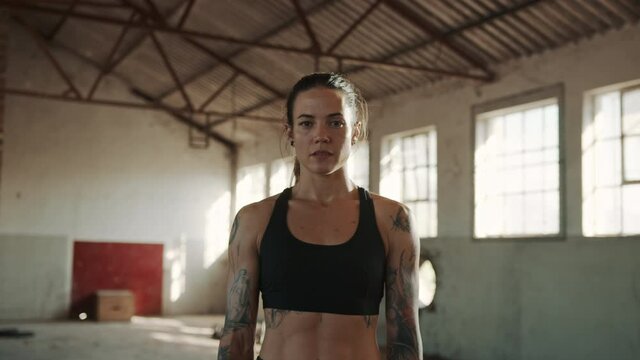 Fit woman standing inside abandoned warehouse after workout. Strong female athlete wearing black sports bra looking at camera.
