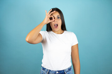 Young beautiful woman standing over isolated blue background doing ok gesture shocked with surprised face, eye looking through fingers