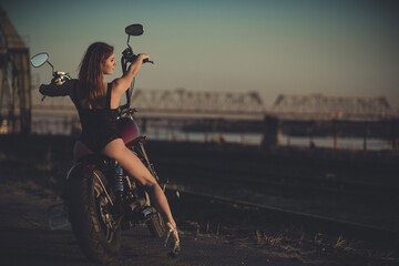 Red-haired woman in sexy lingerie in high heels sits on a motorcycle. Attractive red-haired girl sits on a motorcycle at sunset