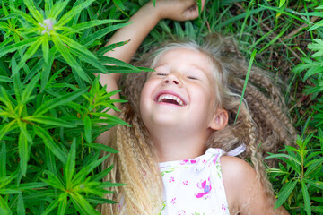 A small Caucasian girl with long blond hair lies on her back in the grass, raised her hand, closed her eyes, laughs. Childhood, happiness, reunion with nature, lifestyle. Close-up portrait of a child