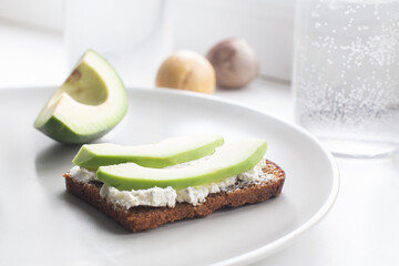 Avocado slices and a sandwich with soft cheese and avocado on a plate, a glass of water. Healthy food concept. Delicious and healthy breakfast. Close-up, selective focus, high key..