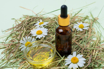 Cosmetic glass brown bottles with dropper, a oil and chamomile flowers on hay on mint background.