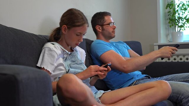 Dad watching TV, daughter plays on smartphone.