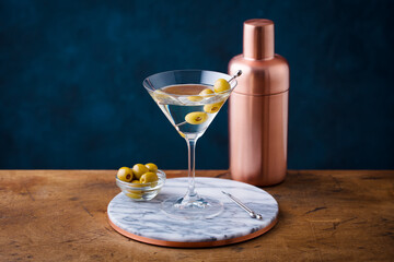 Martini cocktail with green olives, shaker on marble board, wooden table. Copy space.