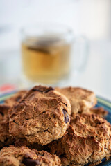 Primer plano de galletas dulces con trozos de chocolate y una taza de infusión (té) de fondo. Colores cálidos y aromas hogareños 
