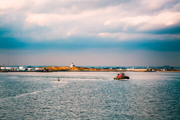 Coast of Bayonne, New Jersey in autumn. Tug boat, industrial buildings and oil storage in New York Harbor, United States. Scenic landscape with dark stormy sky.