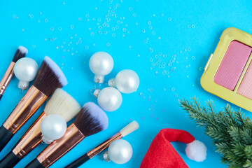 Fototapeta na wymiar creative Christmas cosmetics flat lay - premium makeup brushes, blush palette, eyeshadow palette, Christmas balls, Christmas tree and Santa hat on a colored blue background